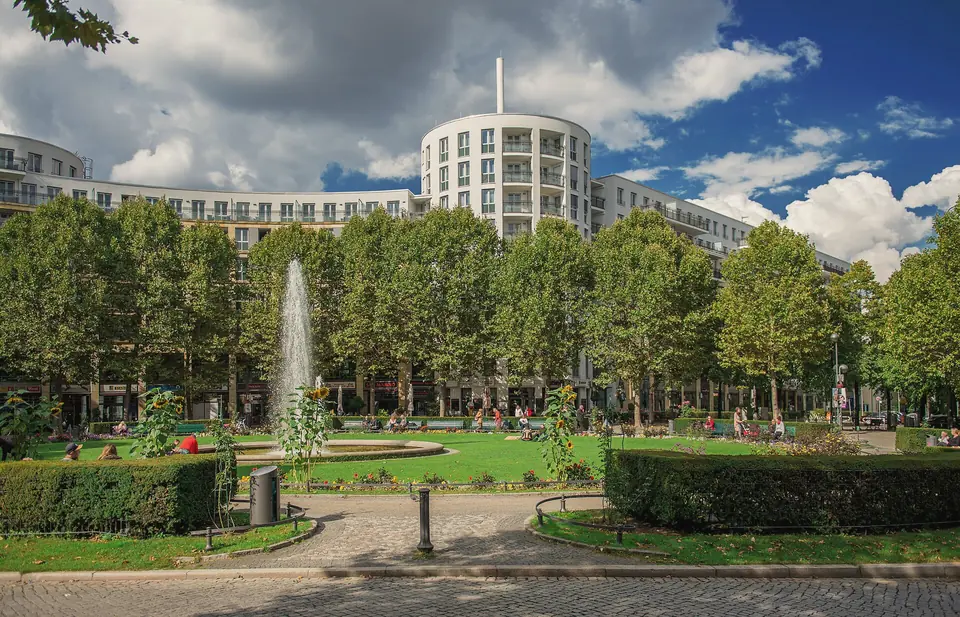 Der Prager Platz mit Springbrunnen, Rasen und Bäumen, dahinter Wohnhäuser der IBA 87