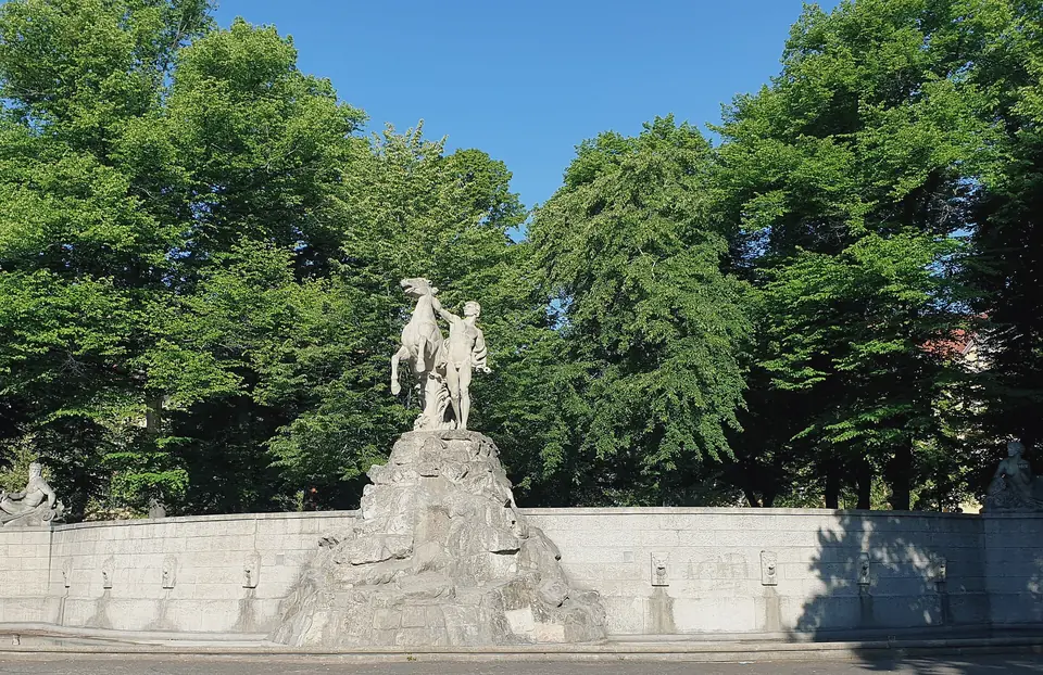 Der Siegfriedbrunnen auf dem Rüdesheimer Platz vor hohen Bäumen unter blauem Himmel