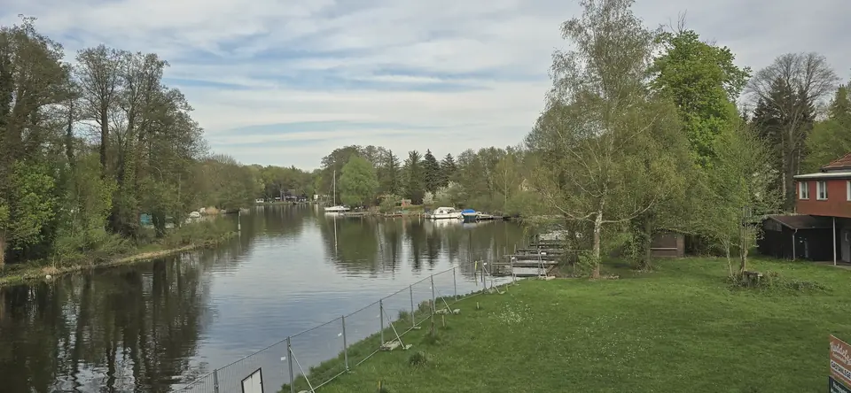Die Dahme zwischen bewaldeten Ufern kurz vor dem Dolgensee, rechts eine Wiese mit Bootsstegen, leicht bewölkter Himmel