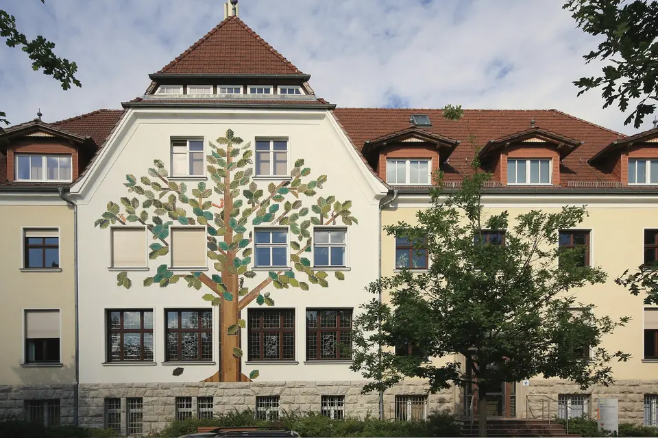 Fassade des Rathauses Eichwalde mit gemaltem Baum über drei Geschossen, rotes Ziegeldach, davor ein Laubbaum