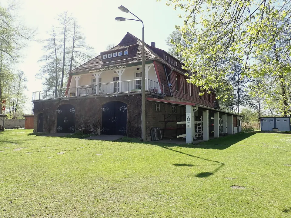 Bootshaus des Ruderclubs Königs Wusterhausen mit Holzbalkon und Bootshallen auf einer Wiese unter Bäumen, sonniger Frühlingstag