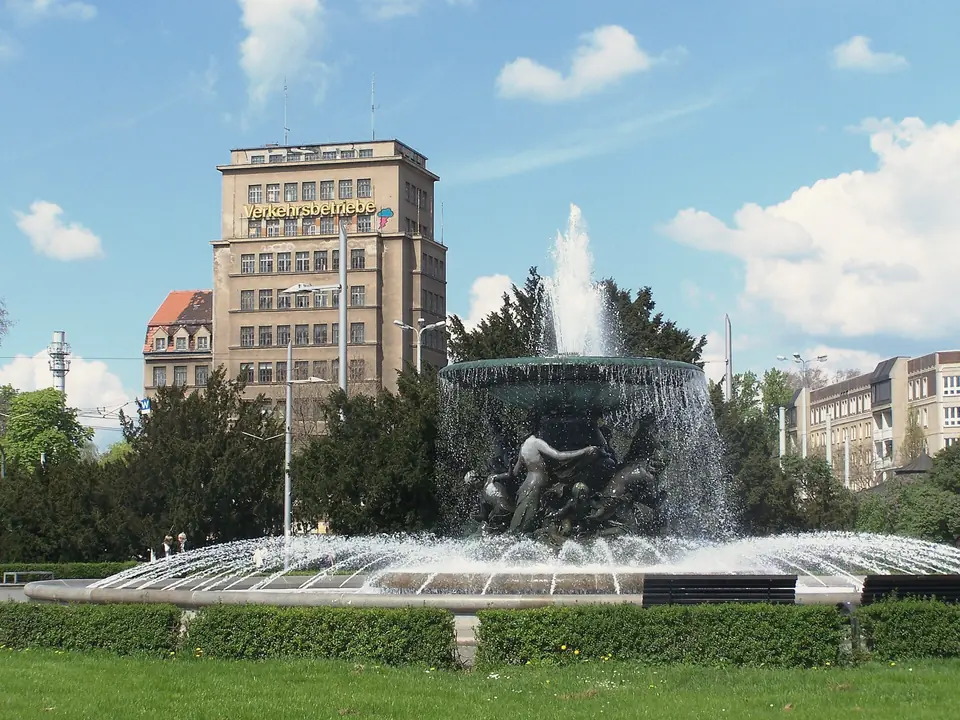 Springbrunnen „Stille Wasser“ auf dem Albertplatz mit Bronzefiguren, dahinter das Hochhaus am Albertplatz und Bäume unter blauem Himmel