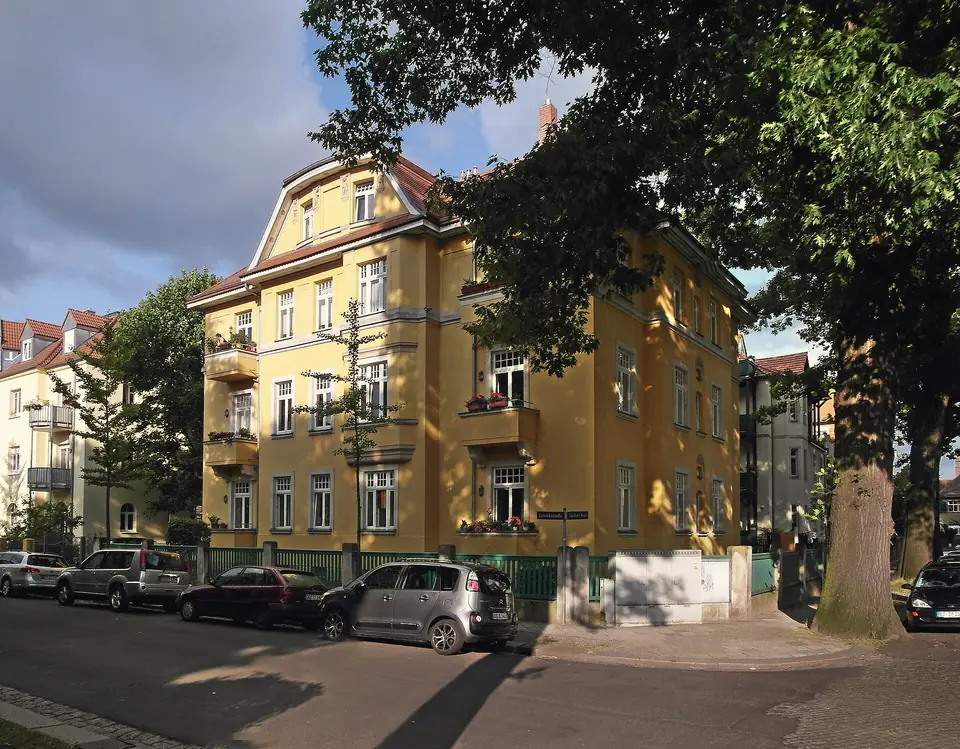 Gelbes viergeschossiges Mietshaus mit Balkonen und Zwerchgiebel an einer Straßenecke in Tolkewitz, davor ein großer Baum und parkende Autos