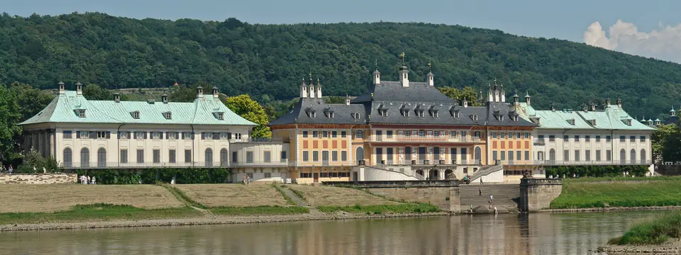 Schloss Pillnitz mit Wasserpalais und Freitreppe am Elbufer, dahinter bewaldete Hänge, im Vordergrund die Elbe