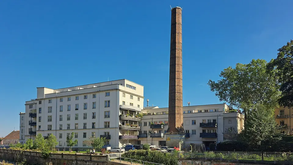 Weiße Gebäude der Bienertmühle in Plauen mit Balkonen und hohem Backsteinschornstein, davor Bäume und Parkplätze, tiefblauer Himmel