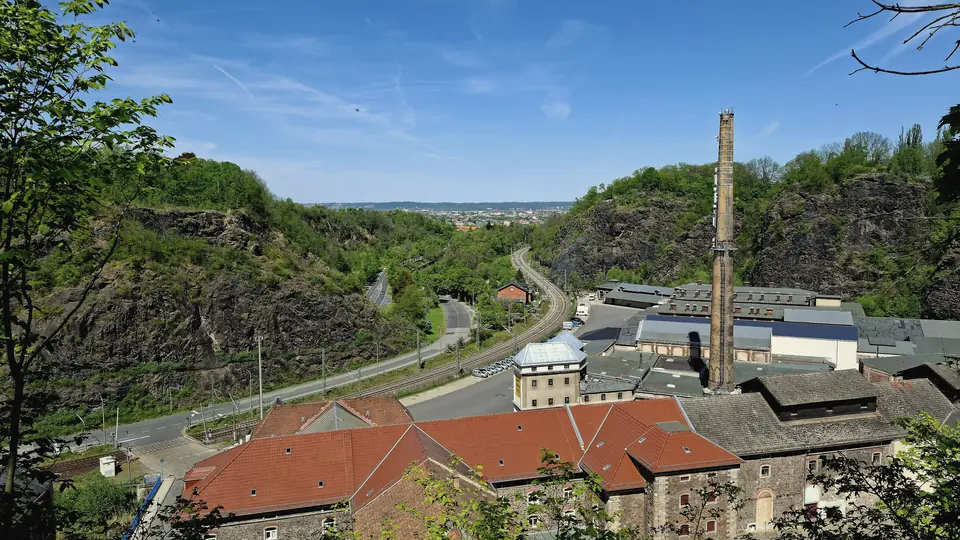 Blick von oben in den Plauenschen Grund mit Felswänden, Bahnstrecke, Straße und einem Fabrikgelände mit Schornstein, dahinter Dresden unter blauem Himmel