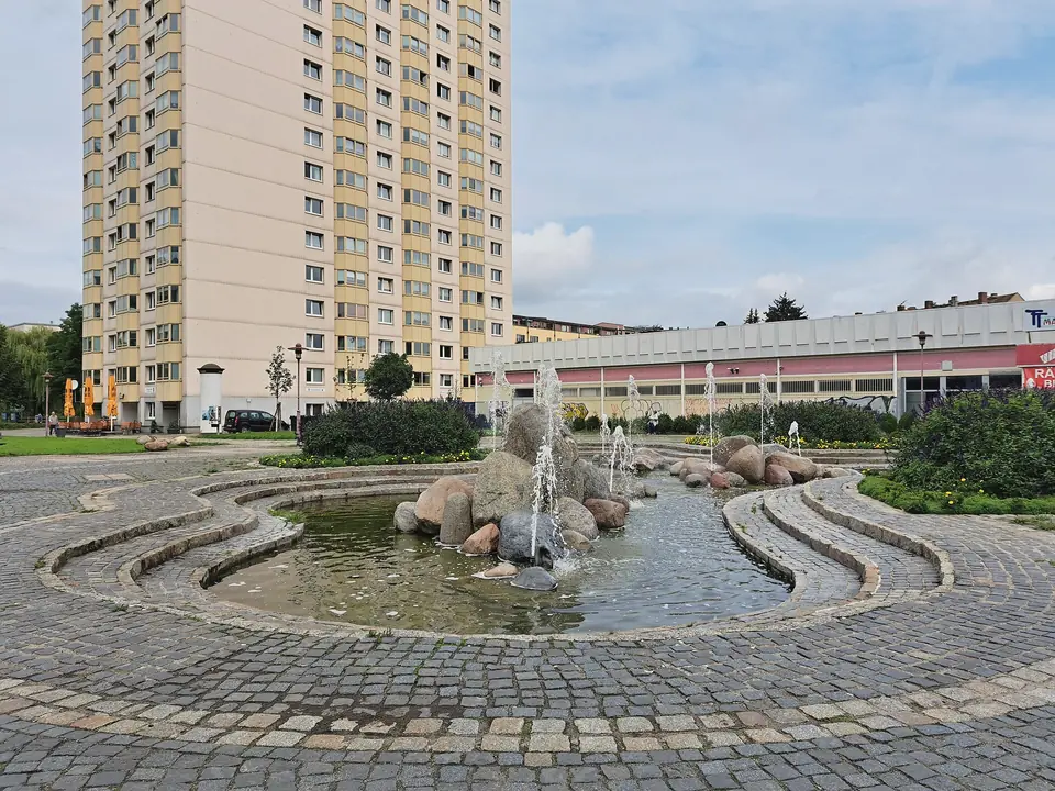 Findlingsbrunnen in Gruna mit Wasserfontänen in einem gepflasterten Becken, dahinter ein Wohnhochhaus und ein flacher Ladenbau