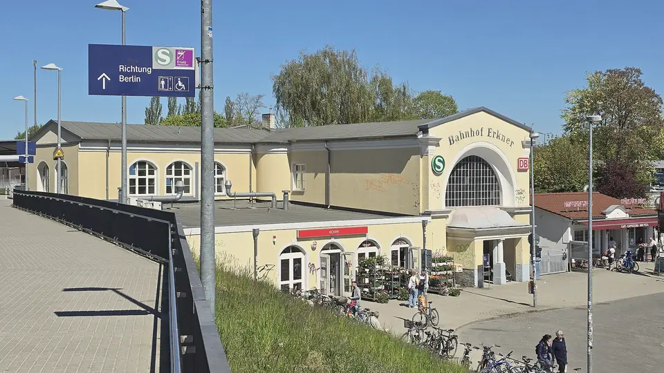 Gelbes Empfangsgebäude des Bahnhofs Erkner mit Rundbogenfenstern, davor Fahrradständer, ein Blumenstand und ein Schild Richtung Berlin, blauer Himmel