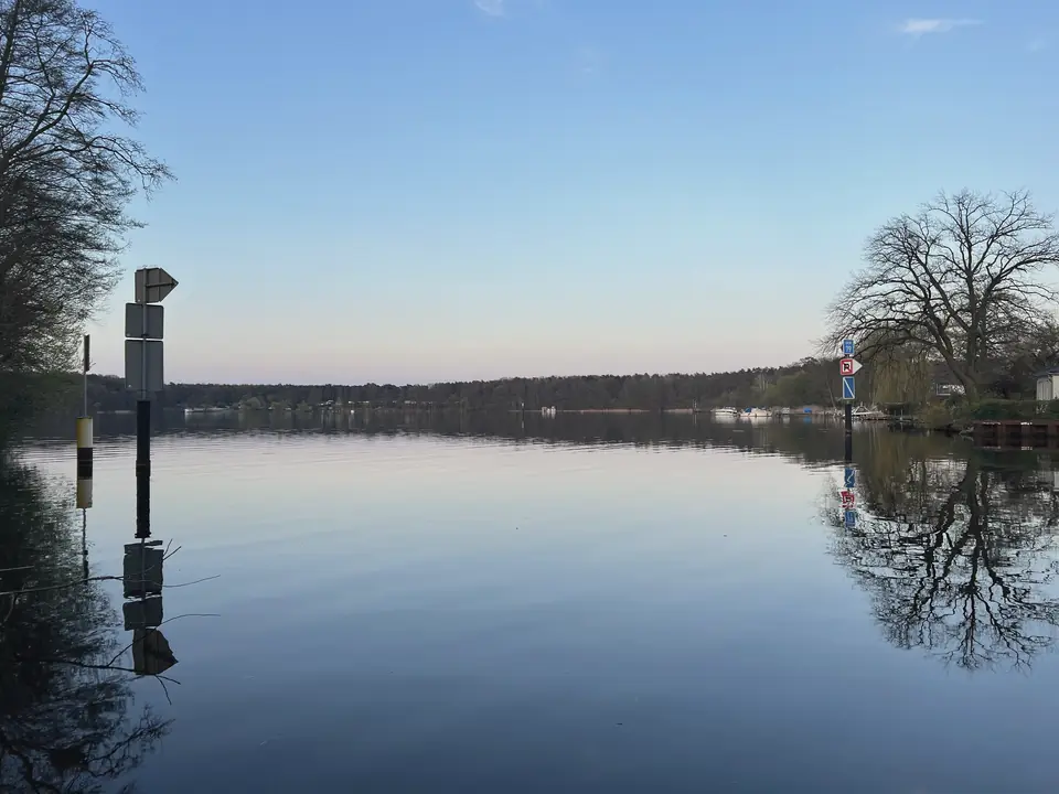 Blick über das Flakenfließ auf den Flakensee mit glattem Wasser, Bäumen am Ufer und Waldrand am Horizont unter klarem Himmel