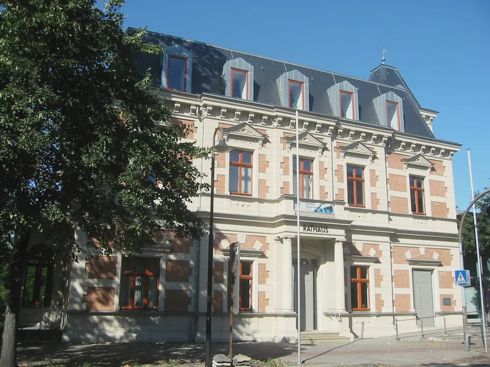Dreigeschossiges Rathaus von Erkner mit heller Fassade, Backsteinflächen, Säulen am Eingang und Mansarddach mit Gauben, links ein großer Laubbaum, blauer Himmel