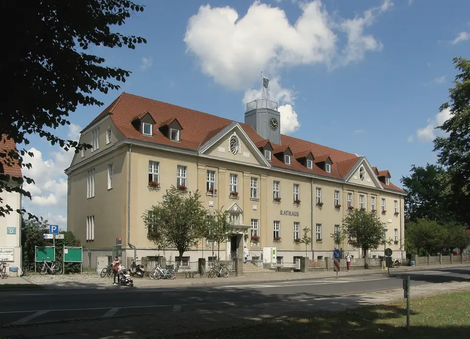 Rathaus Falkensee an der Falkenhagener Straße, dreigeschossiger Bau mit rotem Walmdach, Gauben und Uhrturm, davor Bäume, Fahrräder und die Straße, blauer Himmel mit Wolken