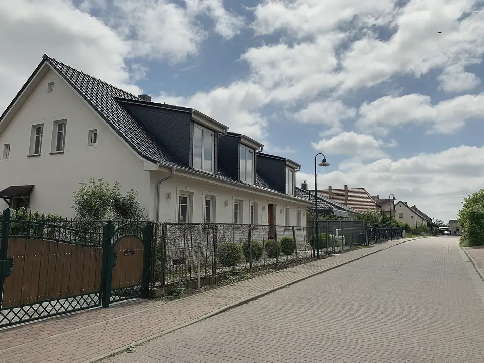Gepflasterte Wohnstraße in Bredow mit einem weißen Einfamilienhaus mit dunklem Dach und Gauben hinter einem Zaun, weitere Häuser dahinter, blauer Himmel mit Wolken