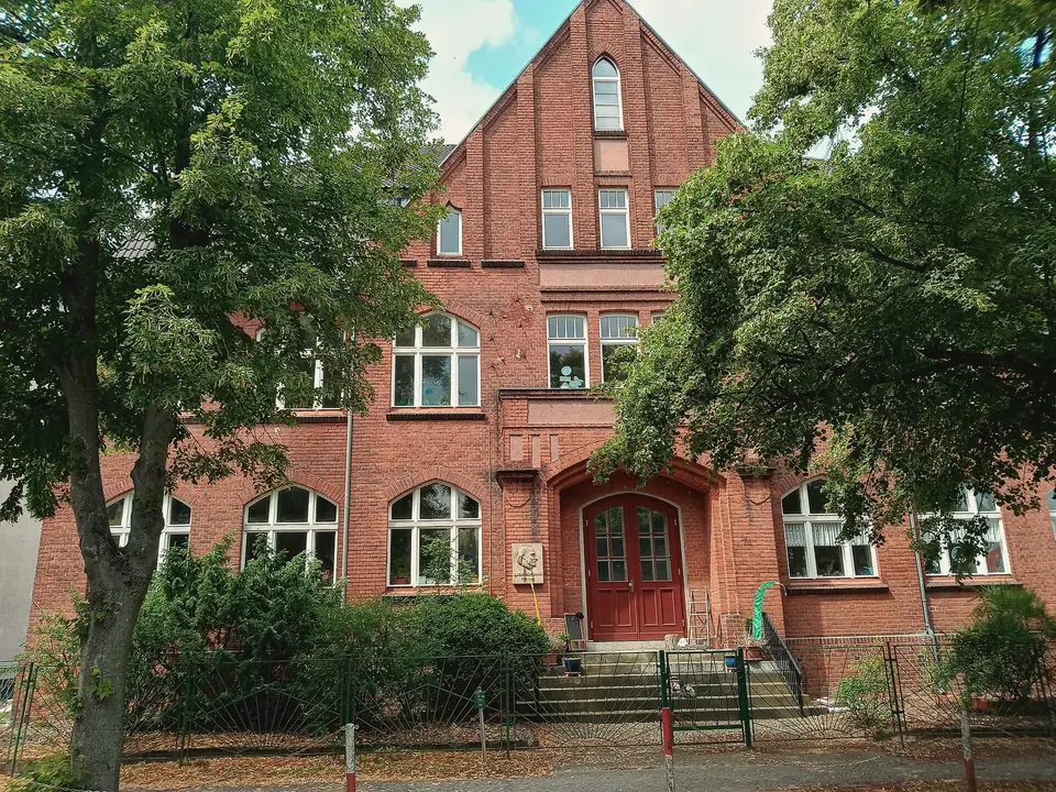 Rote Backsteinfassade der ehemaligen Diesterwegschule in Falkensee mit Spitzgiebel, hohen Fenstern und rotem Portal, davor Laubbäume und ein Zaun
