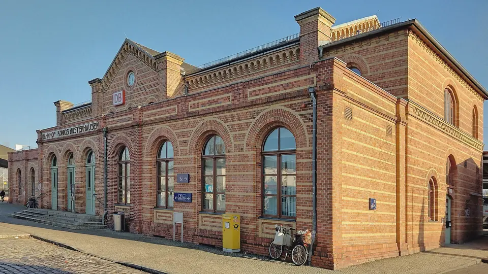 Empfangsgebäude des Bahnhofs Königs Wusterhausen aus gelbem und rotem Backstein mit Rundbogenfenstern und DB-Schild, davor ein gepflasterter Vorplatz mit Fahrrad, blauer Himmel