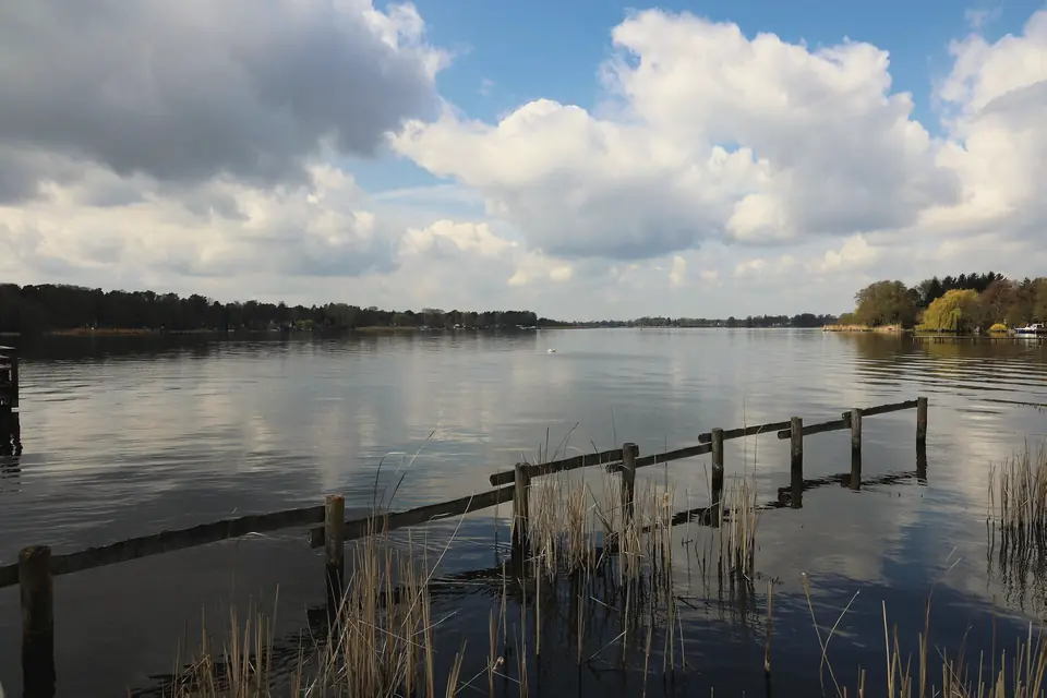 Weiter Blick über den Krüpelsee unter Haufenwolken, vorn Schilf und ein alter Holzzaun im Wasser, am gegenüberliegenden Ufer ein flacher Waldstreifen