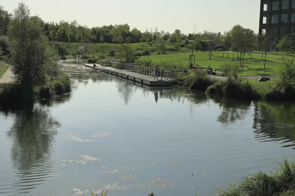Der Karl-Heine-Kanal in einem Bogen mit ruhigem Wasser, am Ufer ein hölzerner Pontonanleger, dahinter Wiesen, Bäume und Sträucher, oben rechts ein dunkles Gebäude, klarer Himmel