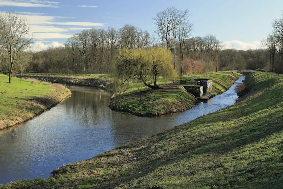 Zwei Wasserläufe zwischen grasbewachsenen Deichen fließen zusammen, auf der Landspitze dazwischen eine Trauerweide und ein kleines Bauwerk am Ufer, dahinter noch kahle Auwaldbäume unter blauem Himmel