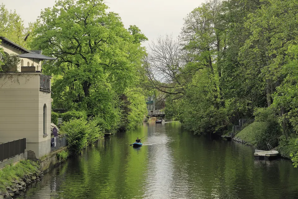 Der Elstermühlgraben zwischen hohen, frisch belaubten Bäumen, links ein helles Haus mit Gitterzaun am Ufer, auf dem Wasser in einiger Entfernung ein Paddelboot, hinten eine Brücke