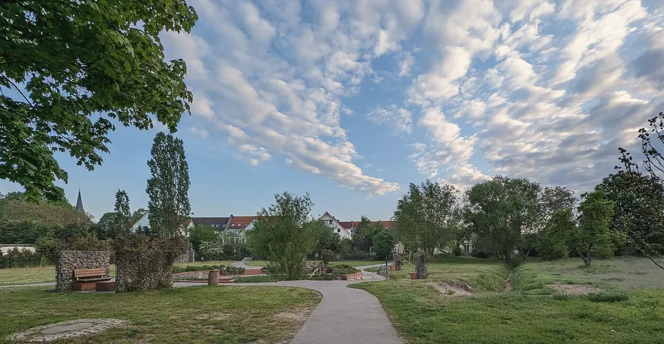 Parkanlage mit geschwungenem Weg, Rasen und jungen Bäumen, hinten Wohnhäuser mit roten Dächern, darüber ein Himmel mit vielen kleinen Wolken