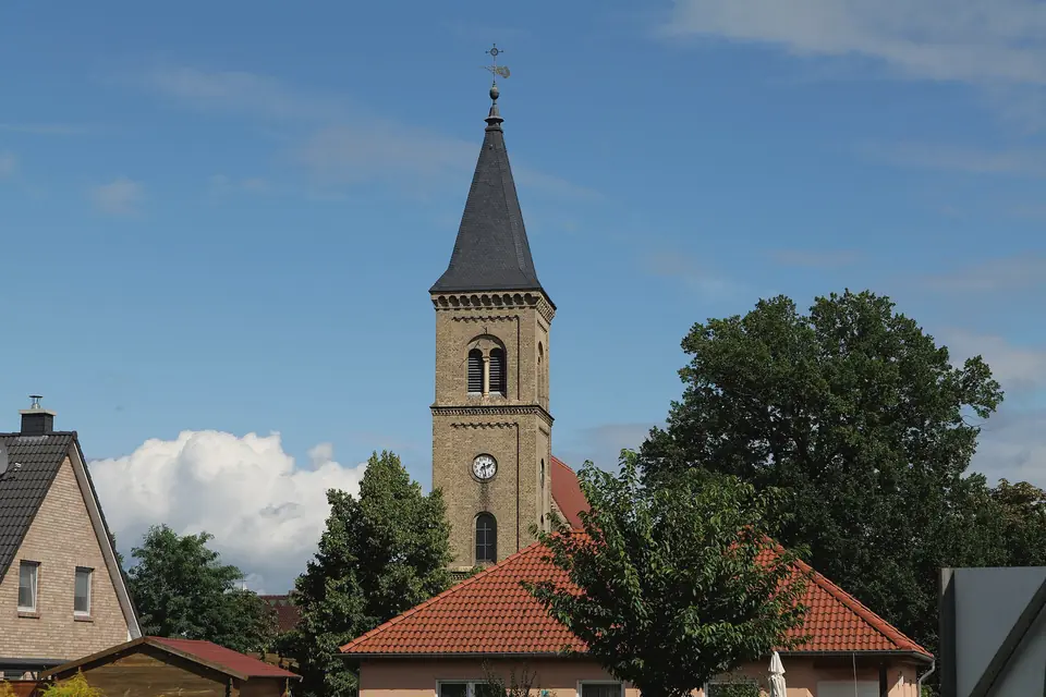 Die Dorfkirche Schönfließ mit spitzem Turm hinter Einfamilienhäusern und Bäumen