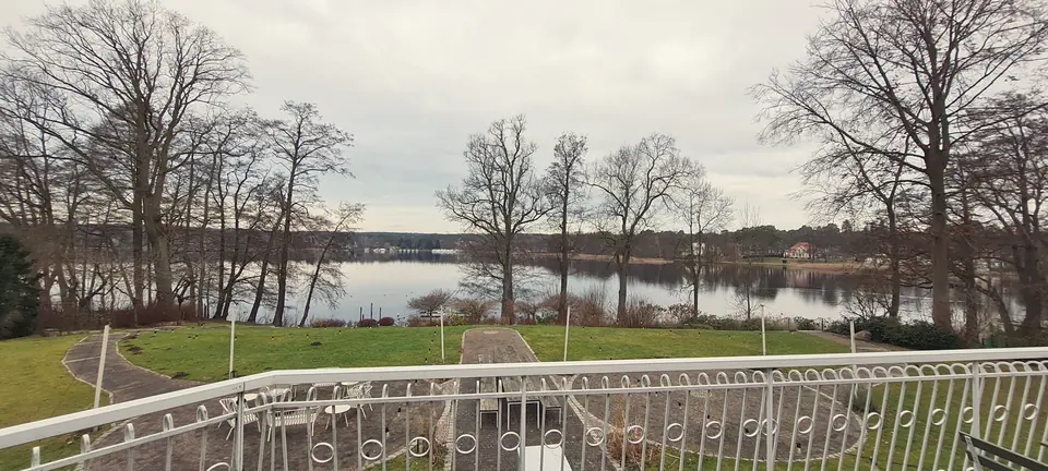 Blick über einen Rasen mit weißem Geländer auf den Scharmützelsee, kahle Bäume am Ufer, gegenüber ein flaches, bewaldetes Ufer unter bedecktem Himmel
