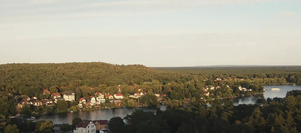 Blick von einer Anhöhe auf Woltersdorf: Häuser mit roten Dächern zwischen Bäumen an einem See, dahinter die bewaldeten Kranichsberge und weites Waldland im Abendlicht