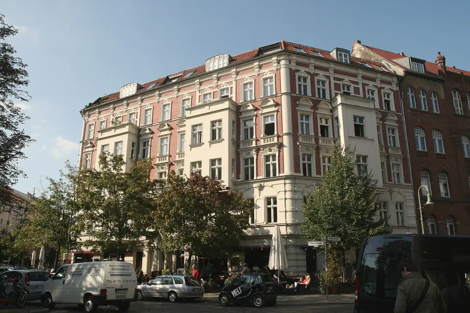 Ein Eckhaus mit rosa und weißer Stuckfassade an der Rykestraße und Knaackstraße, davor Bäume, Cafétische und parkende Autos