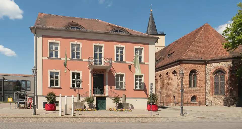 Zweigeschossiges, lachsrotes Rathaus mit Freitreppe und Blumenkübeln, rechts daneben eine Backsteinkirche mit spitzem Turm, blauer Himmel