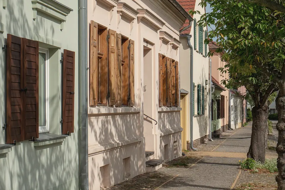 Reihe zweigeschossiger Häuser mit hellen Fassaden und hölzernen Fensterläden an einem gepflasterten Gehweg, rechts Straßenbäume im Sonnenlicht