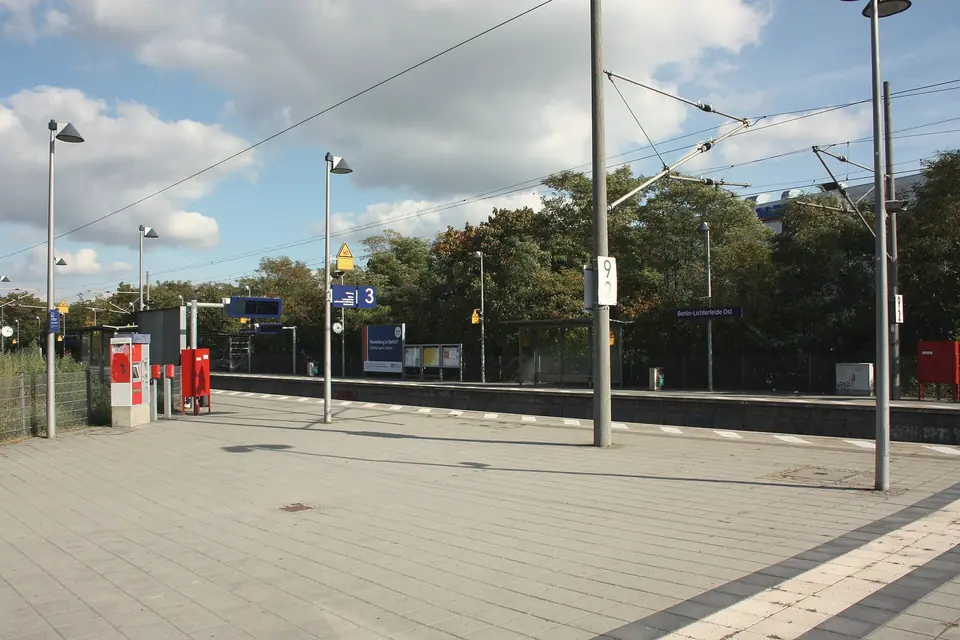Bahnsteig am Bahnhof Lichterfelde Ost mit Bahnsteigschild, Leuchten und Oberleitung unter blauem Himmel mit Wolken