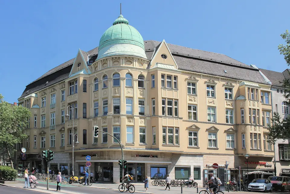 Ein Eckhaus mit grüner Kuppel und heller Fassade an der Schloßstraße in Steglitz, davor Radfahrer und Fußgänger