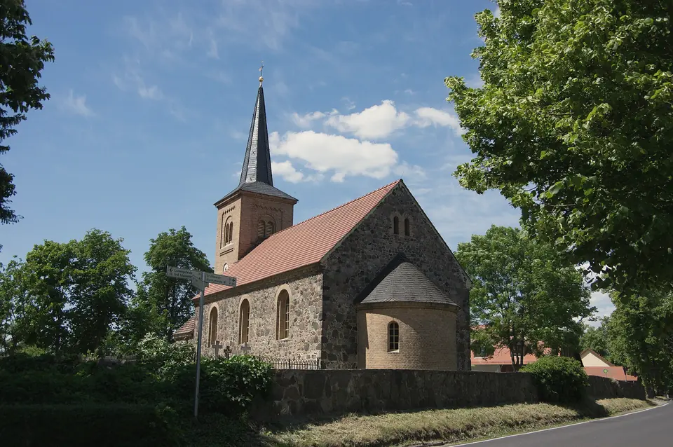 Die Dorfkirche Jühnsdorf aus Feldsteinen mit spitzem Turm und runder Apsis, hinter einer Steinmauer und hohen Laubbäumen