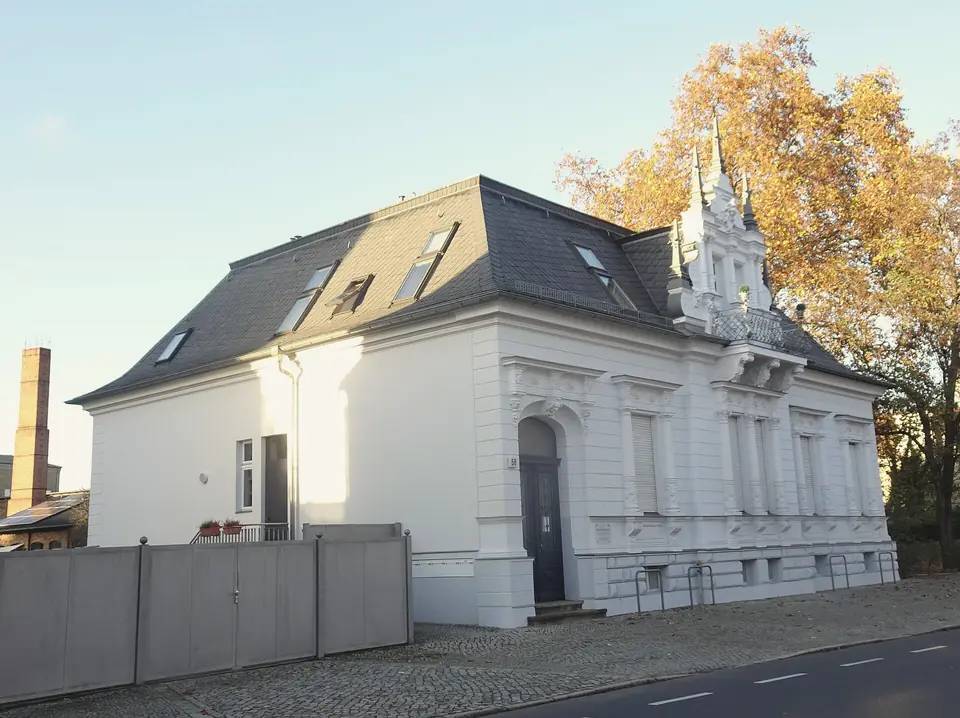 Weiß verputztes Wohnhaus mit Schieferdach, Dachfenstern und verziertem Giebel an einer gepflasterten Straße, dahinter ein Baum in Herbstlaub und ein Backsteinschornstein