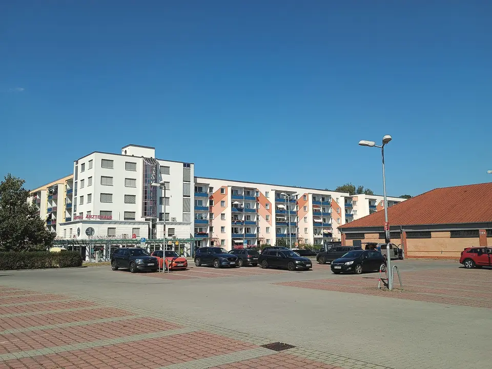 Ein Parkplatz vor hellen Wohn- und Geschäftshäusern mit einem Ärztehaus in Ludwigsfelde unter blauem Himmel