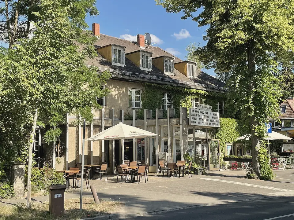 Die Neuen Kammerspiele in Kleinmachnow: ein Haus mit Gauben und Efeu, davor Sonnenschirme und Tische unter hohen Bäumen