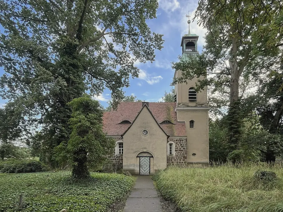 Kleine verputzte Dorfkirche mit Satteldach, rundem Fenster im Giebel und Glockenturm, zwischen hohen Laubbäumen, ein Weg führt durch hohes Gras zur Tür