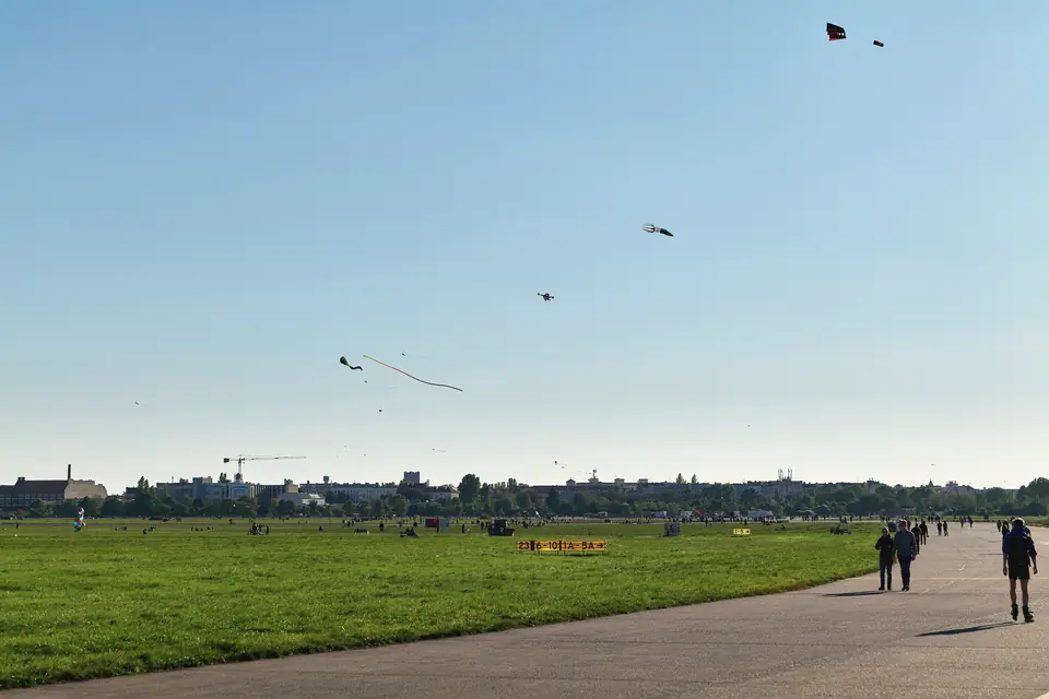 Weite Wiese und Rollbahn auf dem Tempelhofer Feld unter blauem Himmel, am Himmel Drachen, am Horizont die Stadt